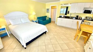 Une chambre à coucher comprenant un lit, des tables de chevet, une commode, un canapé bleu, une chaise en bois et une kitchenette équipée de placards et d’appareils électroménagers.