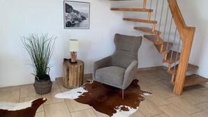 Ein modernes Wohnzimmer mit einem grauen Sessel, Holzboden, einem Kuhfellteppich, einem Holzschemel mit Lampe, einer Topfpflanze und einem gerahmten Foto an der Wand.