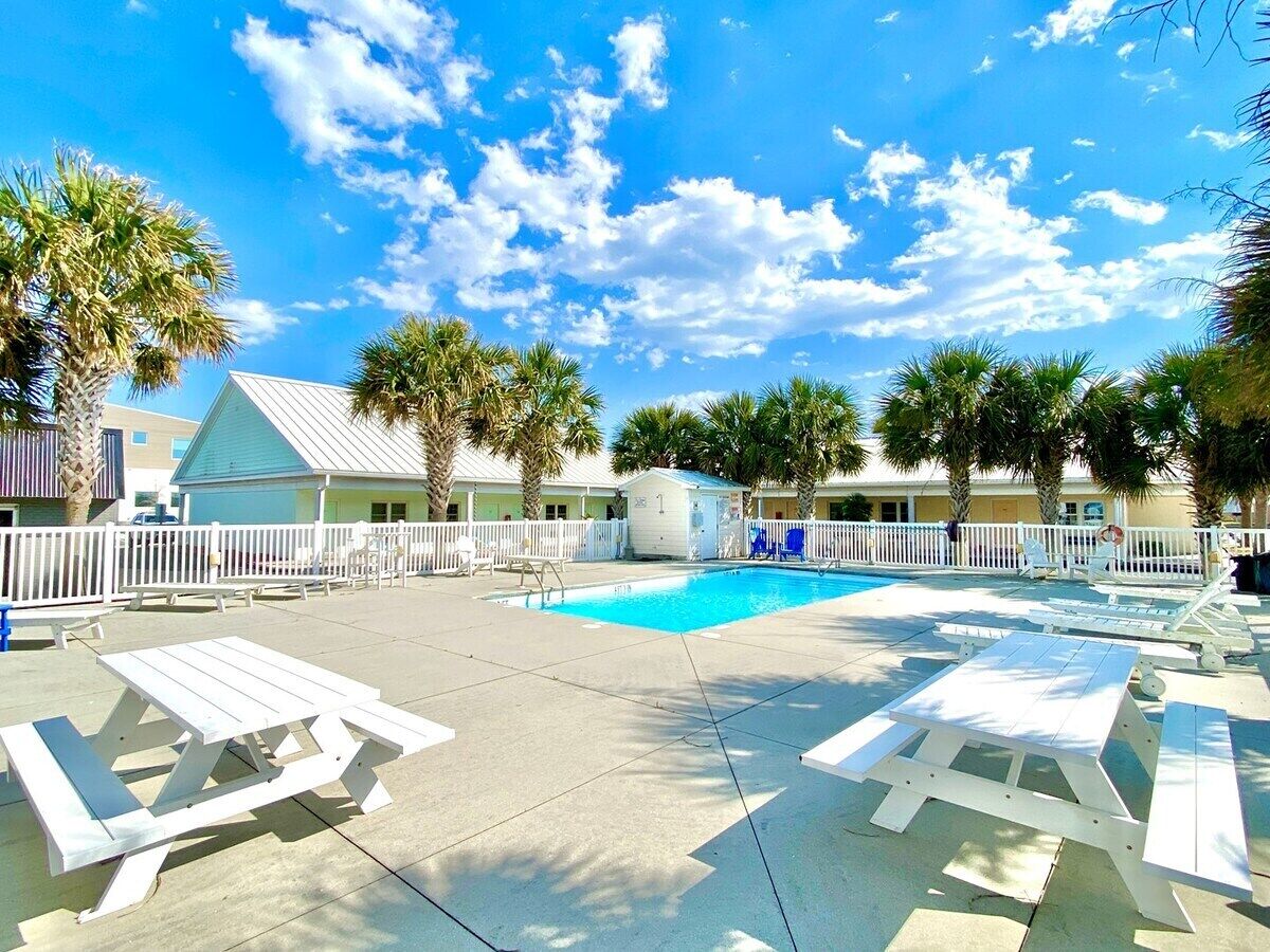 A pool area with white picnic tables and lounge chairs, surrounded by palm trees and a white fence.