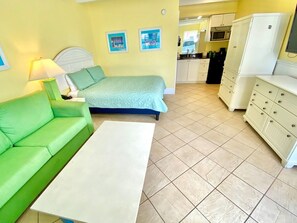 A room with a green sofa, a bed with a blue bedspread, a white table, and a kitchen area with cabinets and a microwave.