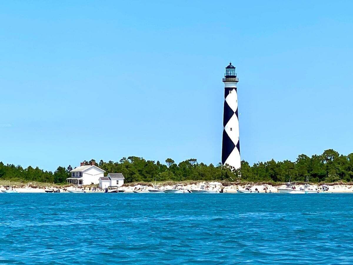 A lighthouse with a white and black pattern, a beach with white sand, and a few boats.