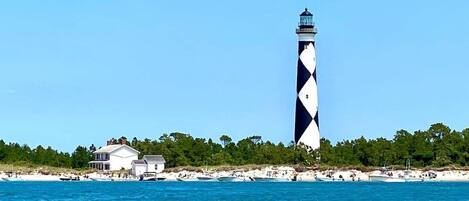 A lighthouse with a white and black pattern, a beach with white sand, and a few boats.