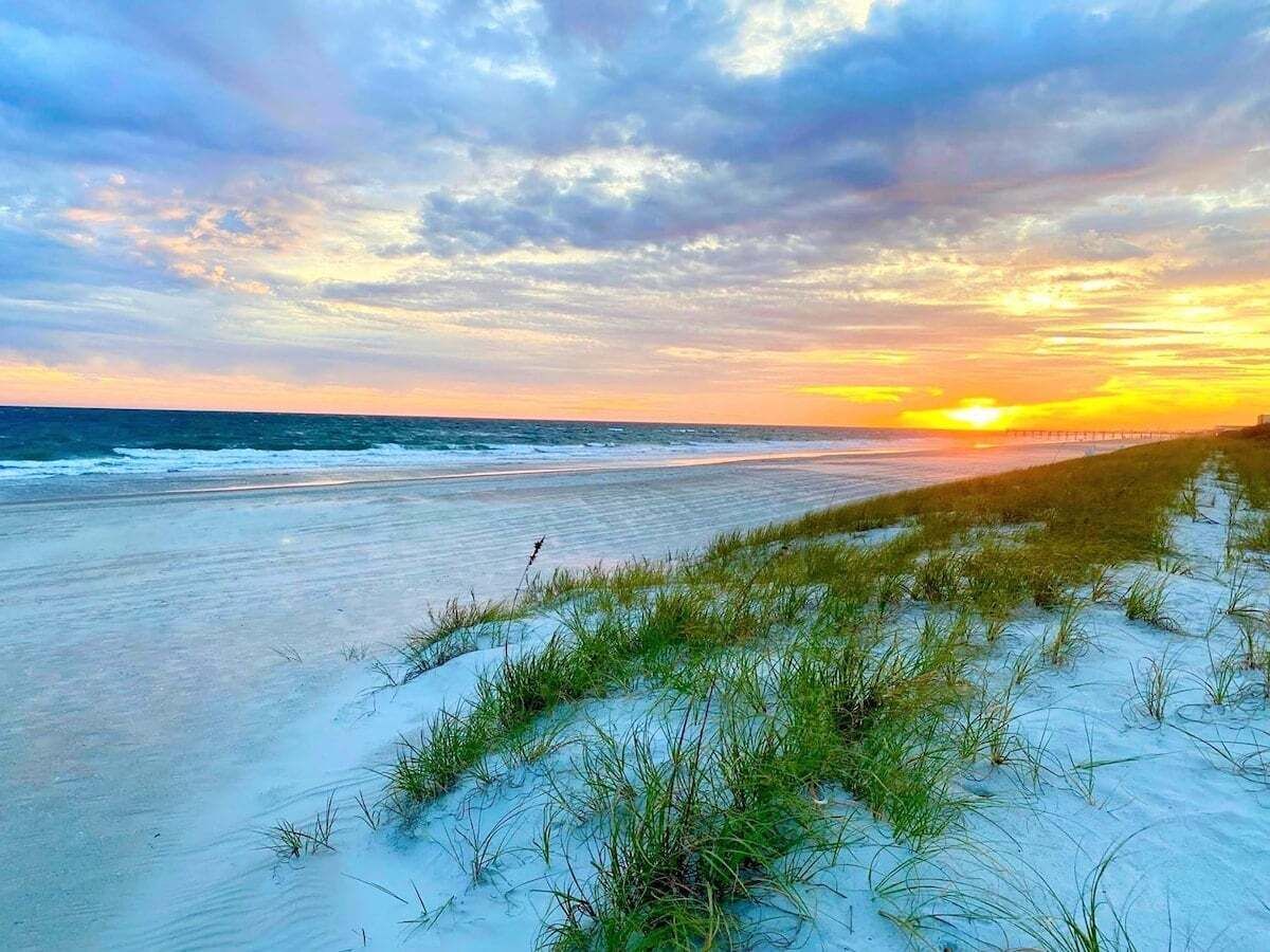 Une plage avec des dunes de sable et de l’herbe, un coucher de soleil en arrière-plan et une jetée qui s’avance dans l’eau.