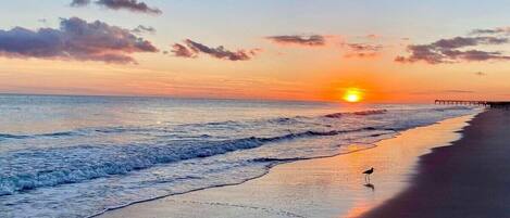 A beach scene with a sunset, ocean waves, and a pier in the distance.