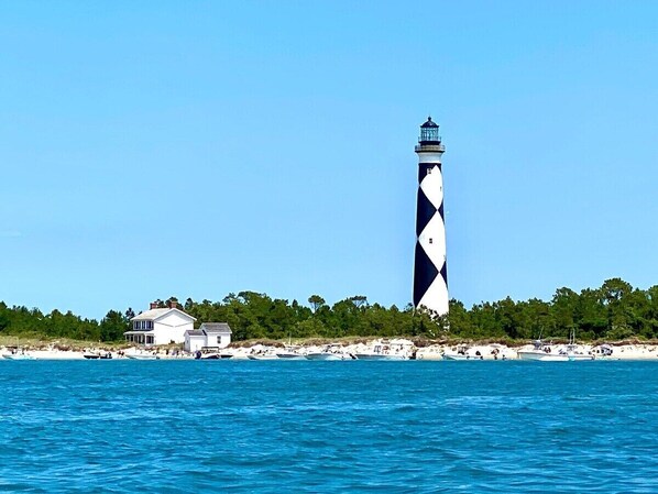 Un phare aux motifs noirs et blancs, un bâtiment blanc doté d’un balcon, et une plage avec des bateaux et des gens.