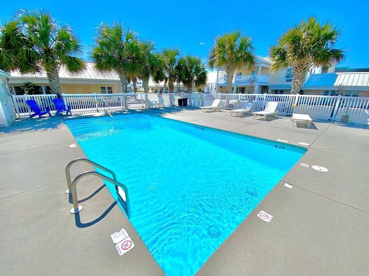A swimming pool with a white fence, lounge chairs, and palm trees.