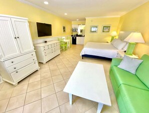 Een kamer met een bed, een groene bank, een witte kapstok, een witte tafel en een kitchenette.