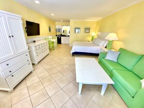A room with a bed, a green sofa, a white dresser, a white table, and a kitchen area in the background.
