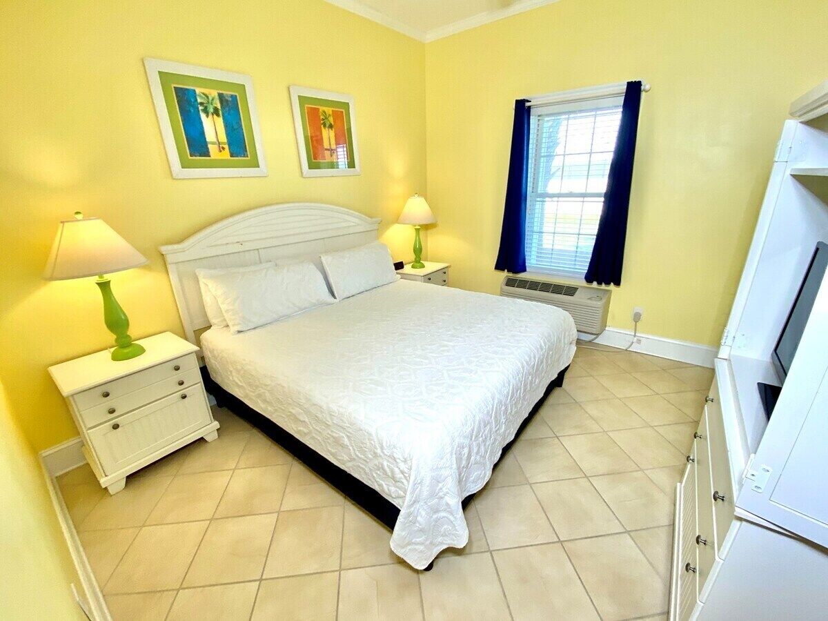 Un dormitorio con cama, dos mesitas de noche, una ventana con cortinas y un aire acondicionado de pared.