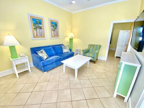 A living room with a blue sofa, white coffee table, green armchair, and a TV stand with cabinets. - Pet-friendly, 3 Blocks to Beach, Pool, #501 (Atlantic Beach)