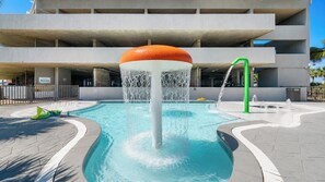 A swimming pool with a central water feature, surrounded by a paved area and a multi-story building in the background. - Beautiful 3-Bedroom with a Balcony - Seawind 905 (Gulf Shores)