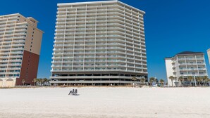 Un hotel frente a la playa con varios pisos, balcones y una playa arenosa al frente.