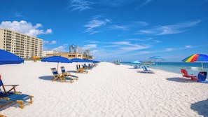 A beach with rows of sun loungers and umbrellas, a hotel building, and clear blue skies. - Beautiful 3-Bedroom with a Balcony - Seawind 905 (Gulf Shores)
