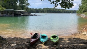 A lakeside view with kayaks on the shore, a covered dock, and a forested backdrop. - 2 BR Hidden Gem with Lake Access, Pool & Hot Tub 15 Min from Lanier Islands  (FLOWERY BRANCH)