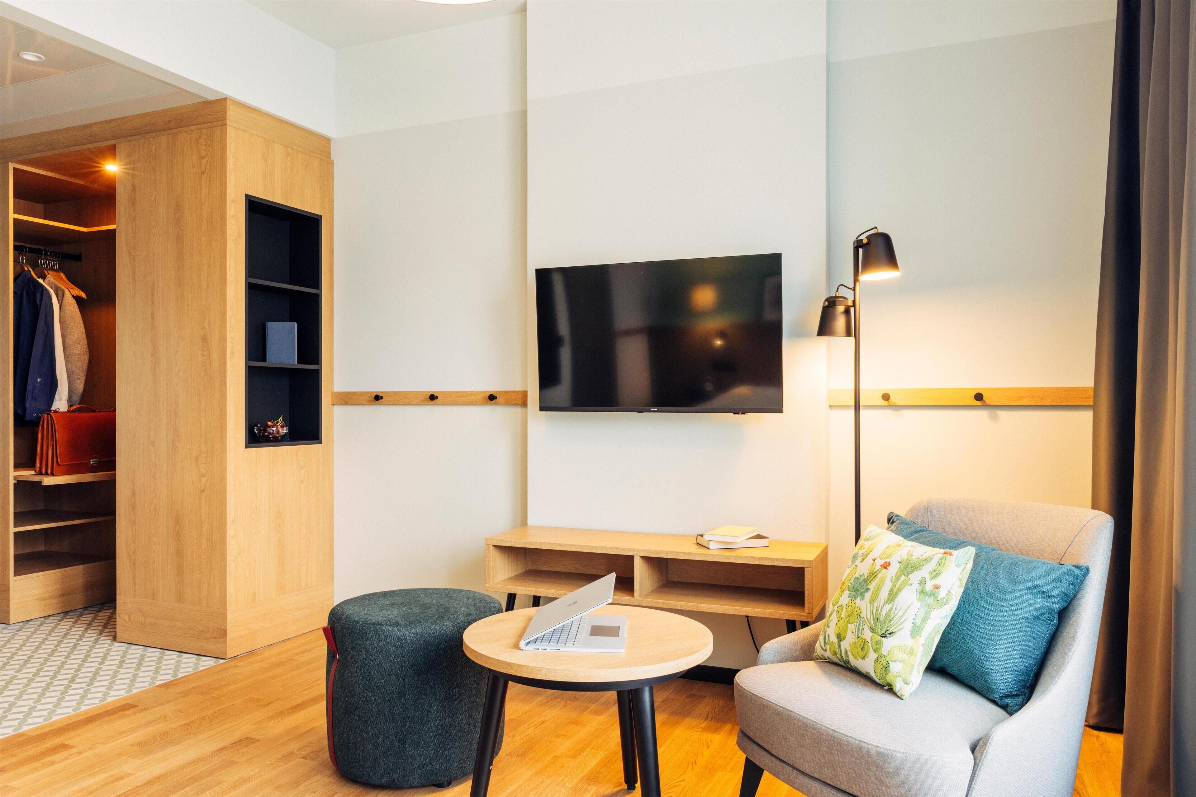 A modern hotel room with a wooden wardrobe, a TV mounted on the wall, a desk with a laptop, a grey armchair, and a small round table.