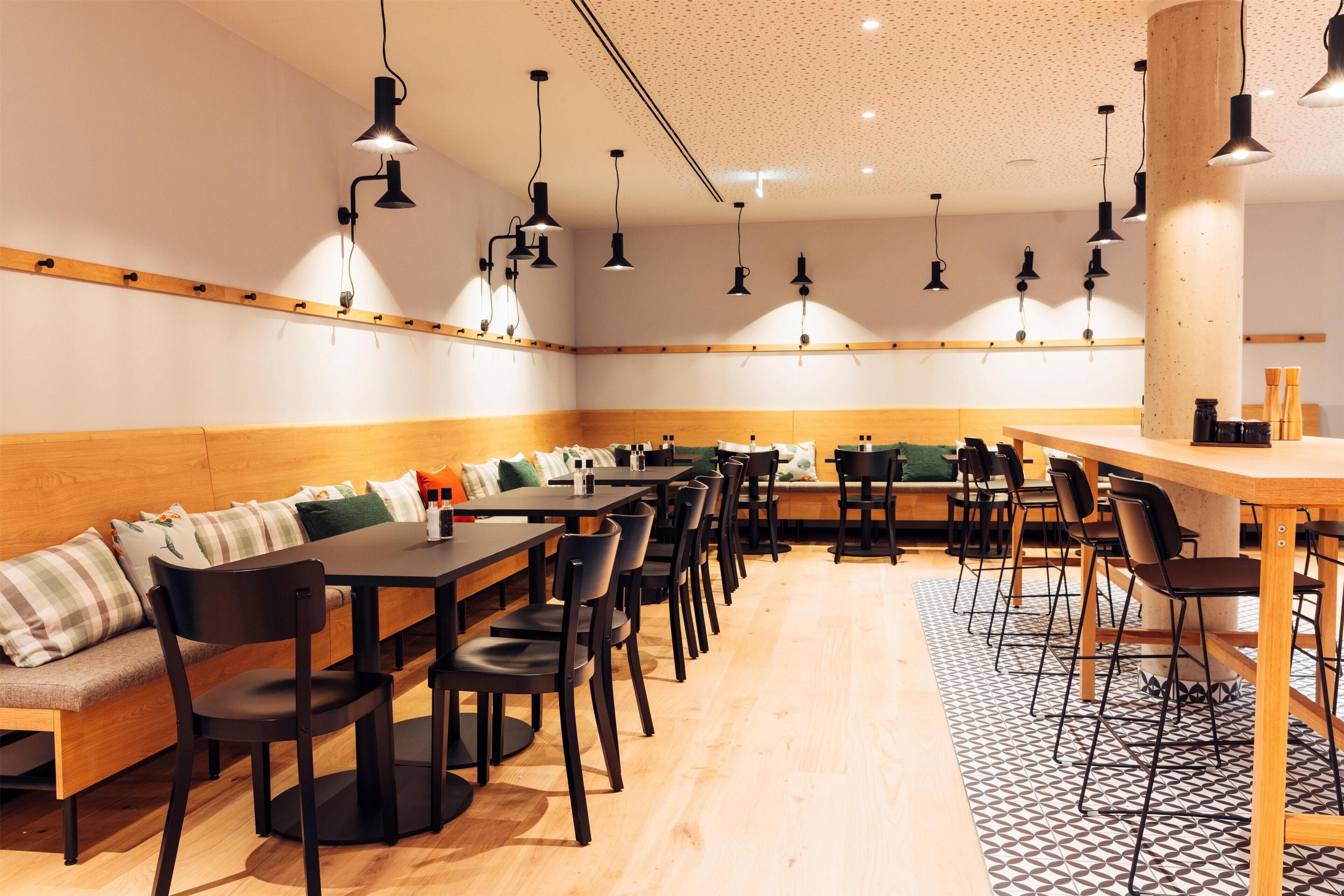 Un restaurante moderno con mesas y sillas de madera, una zona de estar con un largo banco y lámparas colgantes.