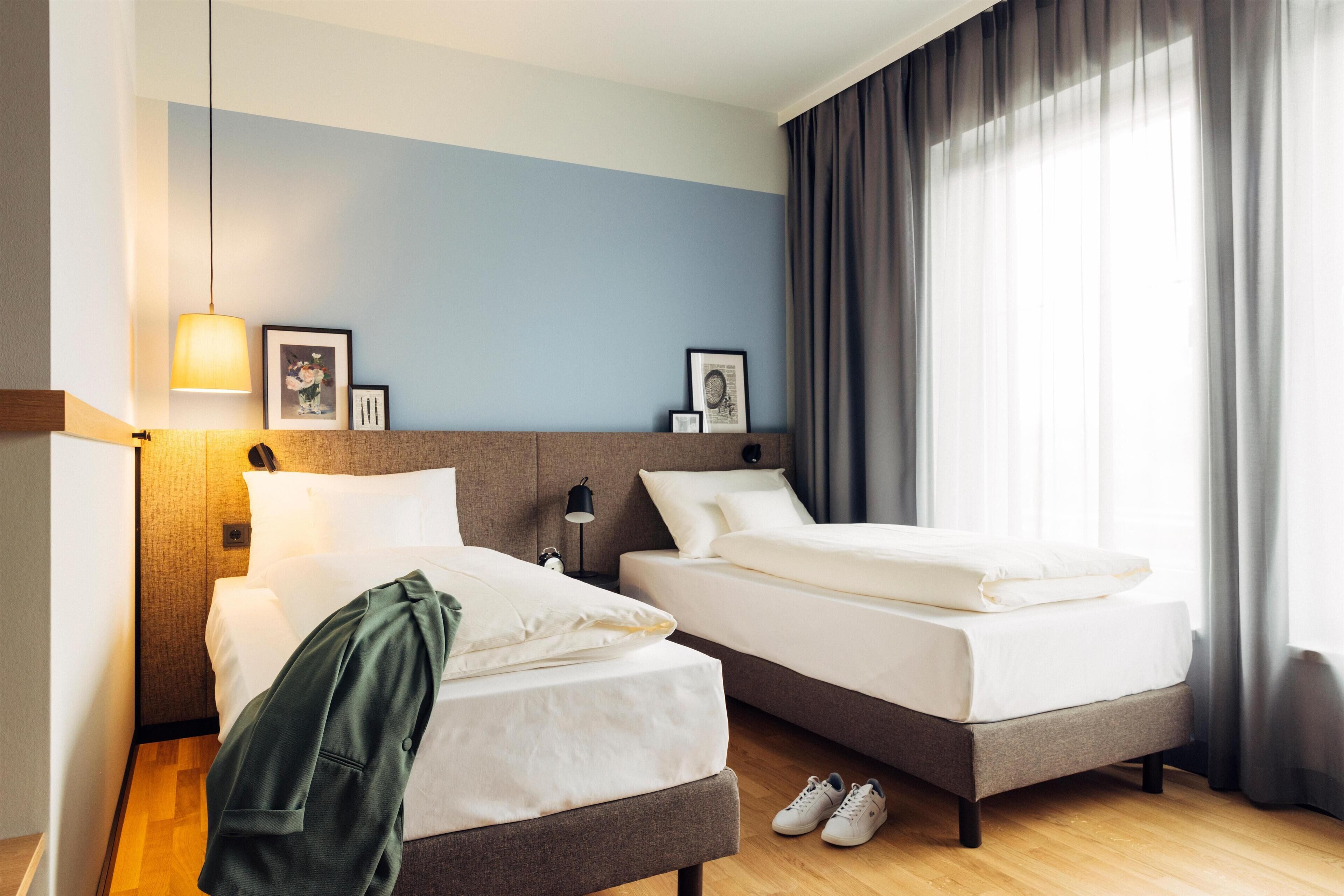 Ein Hotelzimmer mit zwei Betten, einer Lampe, gerahmten Bildern an der Wand und einem Fenster mit Vorhängen.