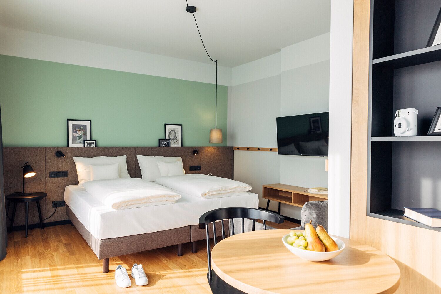Quarto de hotel com duas camas, uma mesa, uma cadeira, televisão e uma estante com câmera e livros.