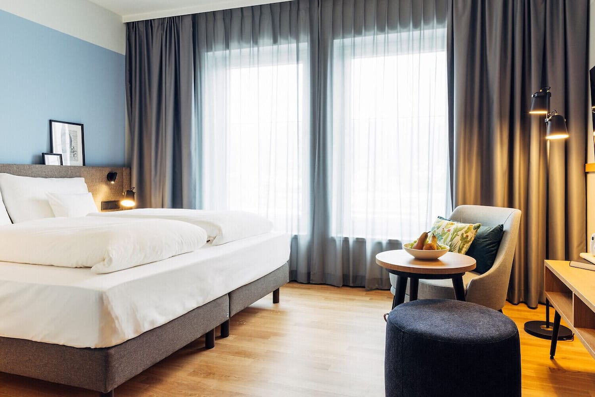 Habitación de hotel con una cama grande, una mesa redonda con un frutero, una silla y un escritorio con una lámpara.