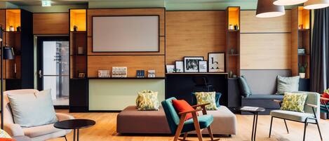 A modern hotel lobby with a wooden reception desk, seating area with sofas and chairs, and a large wall-mounted screen.