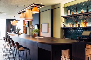 Un espace bar moderne doté d’un long comptoir, de nombreux tabourets, d’un éclairage suspendu et d’une étagère où sont exposées des bouteilles d’alcool.