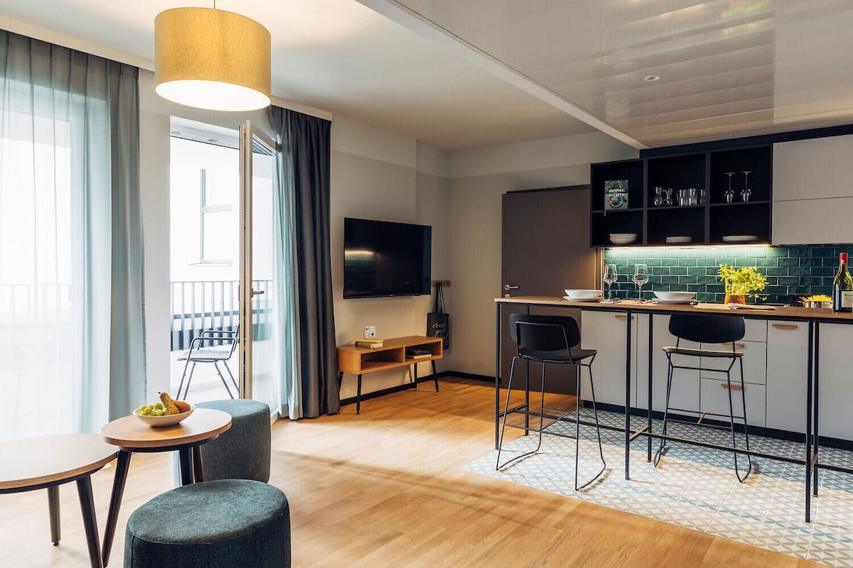 Una sala moderna con piso de madera, una mesa de comedor y dos sillas. Hay una zona de cocina con una barra, taburetes y un televisor montado arriba.