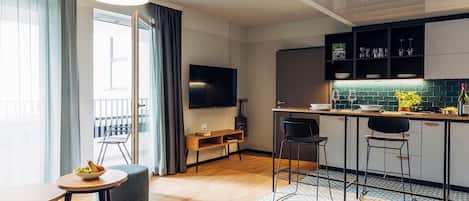 A modern living room with a wooden floor, a dining table, and two chairs. There is a kitchen area with a bar counter, stools, and a TV mounted above it.