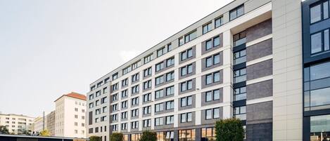 Un hotel moderno de varios pisos con fachada plana, ventanas amplias y un patio central con zonas de estar y jardineras.