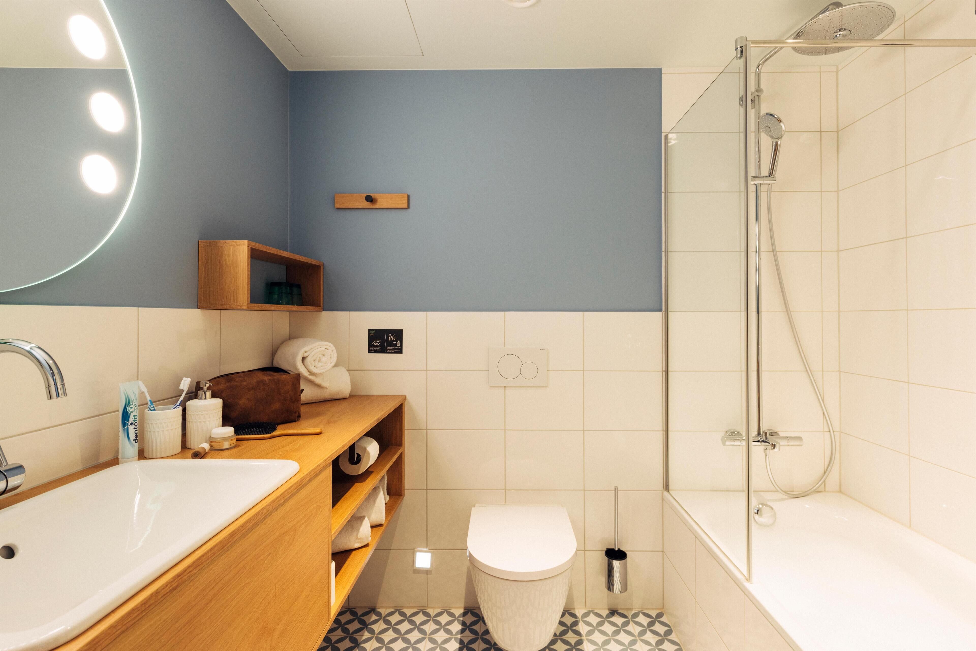 A bathroom with a sink, toilet, and shower. There is a mirror with lights, a shelf with toiletries, and a towel rack.