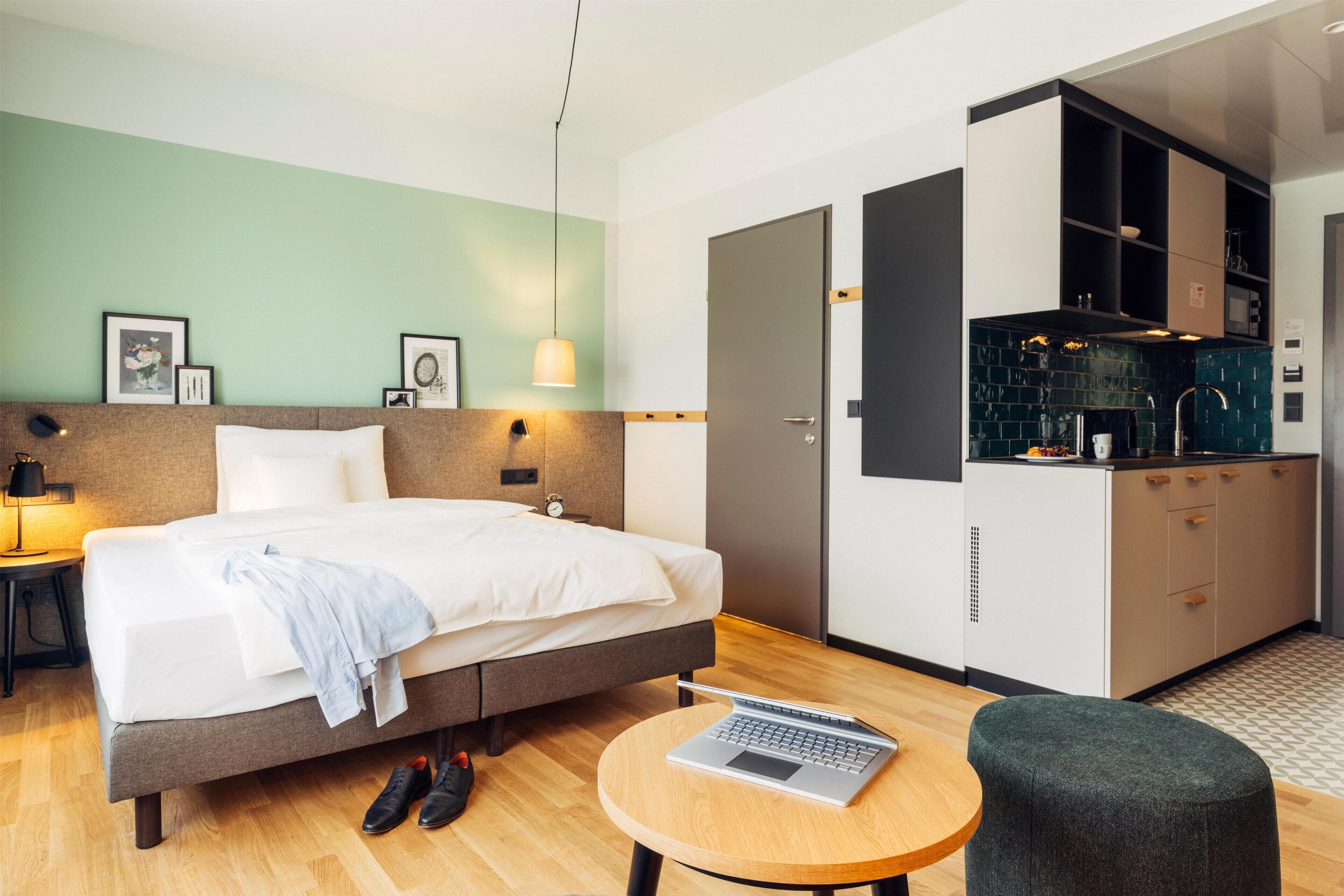 A hotel room with a bed, a laptop on a small table, a shoe rack, and a kitchenette.