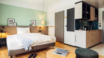 Habitación de hotel con cama, una laptop sobre una mesita, zapatero y kitchenette.