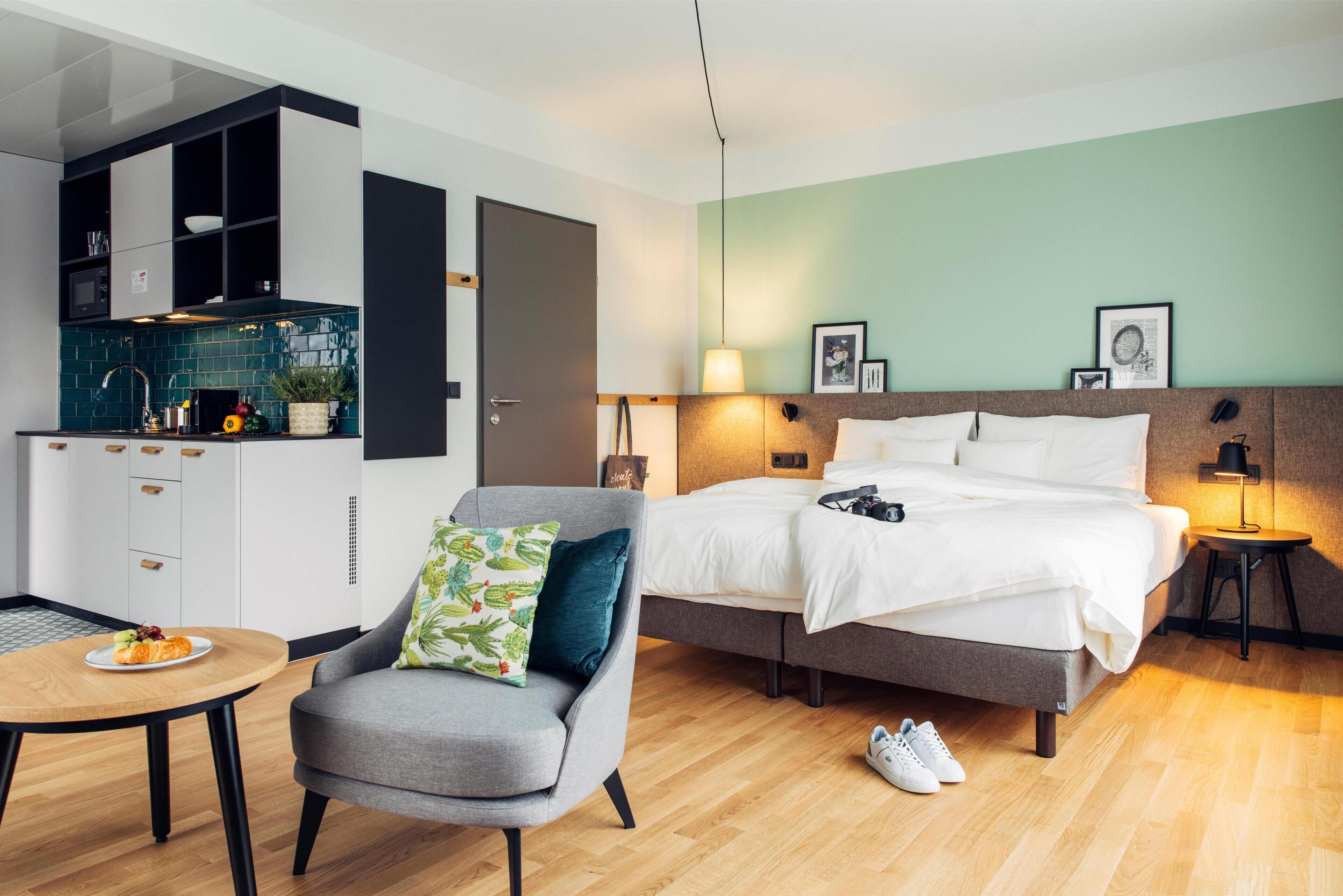 A modern hotel room with a bed, a small desk, a chair, a table with a plate of food, and a kitchenette.