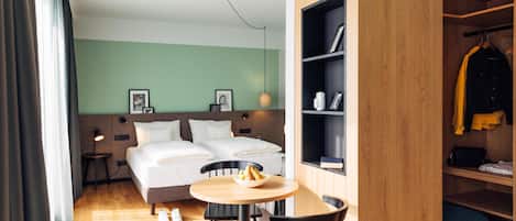 A hotel room with a bed, a round wooden table with two chairs, a small side table, a wardrobe, and a hanging coat rack.