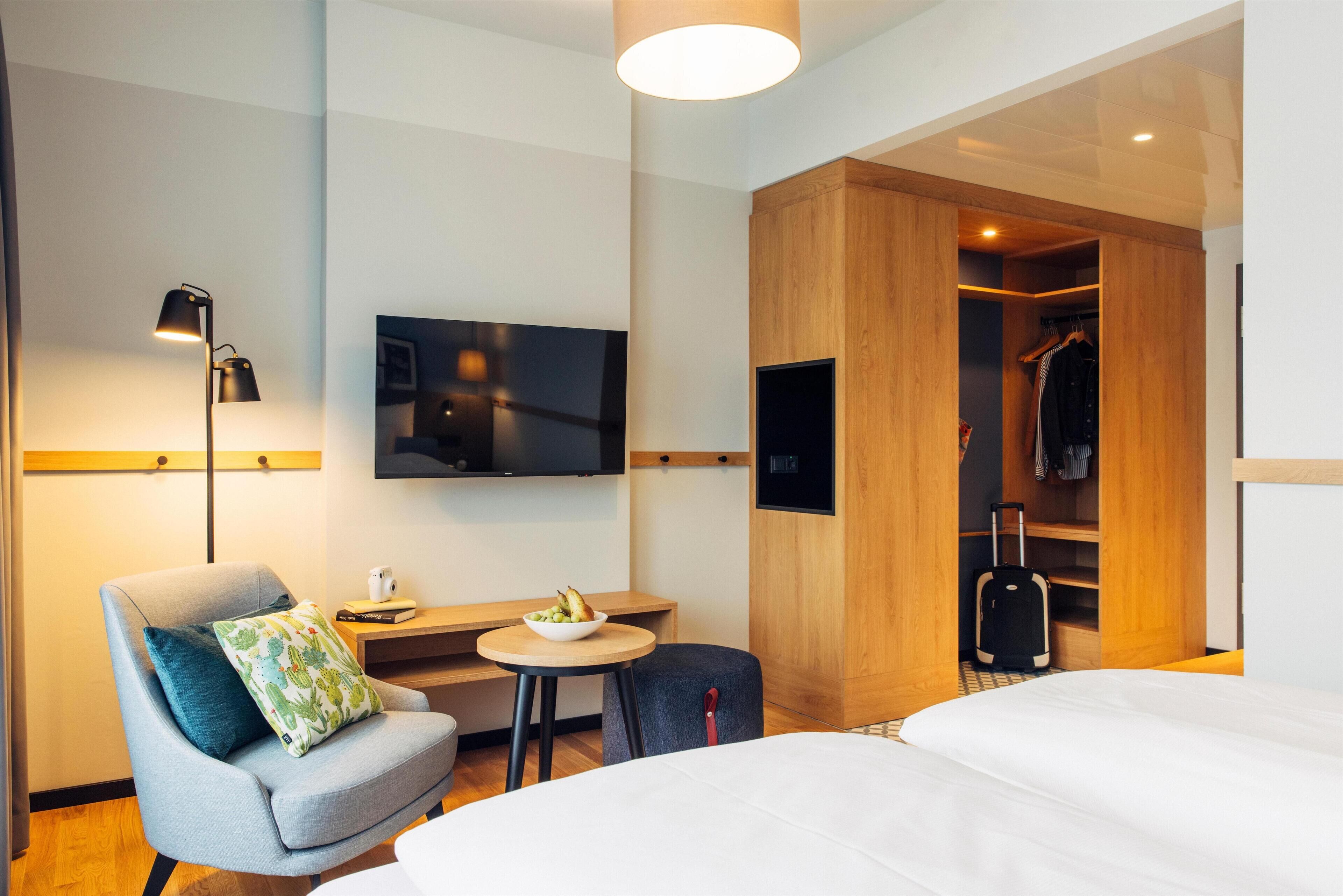 Ein modernes Hotelzimmer mit einem Bett, einem Schreibtisch mit Obstschale, einem Sessel, einem Fernseher, einem Kleiderschrank und einem Koffer.