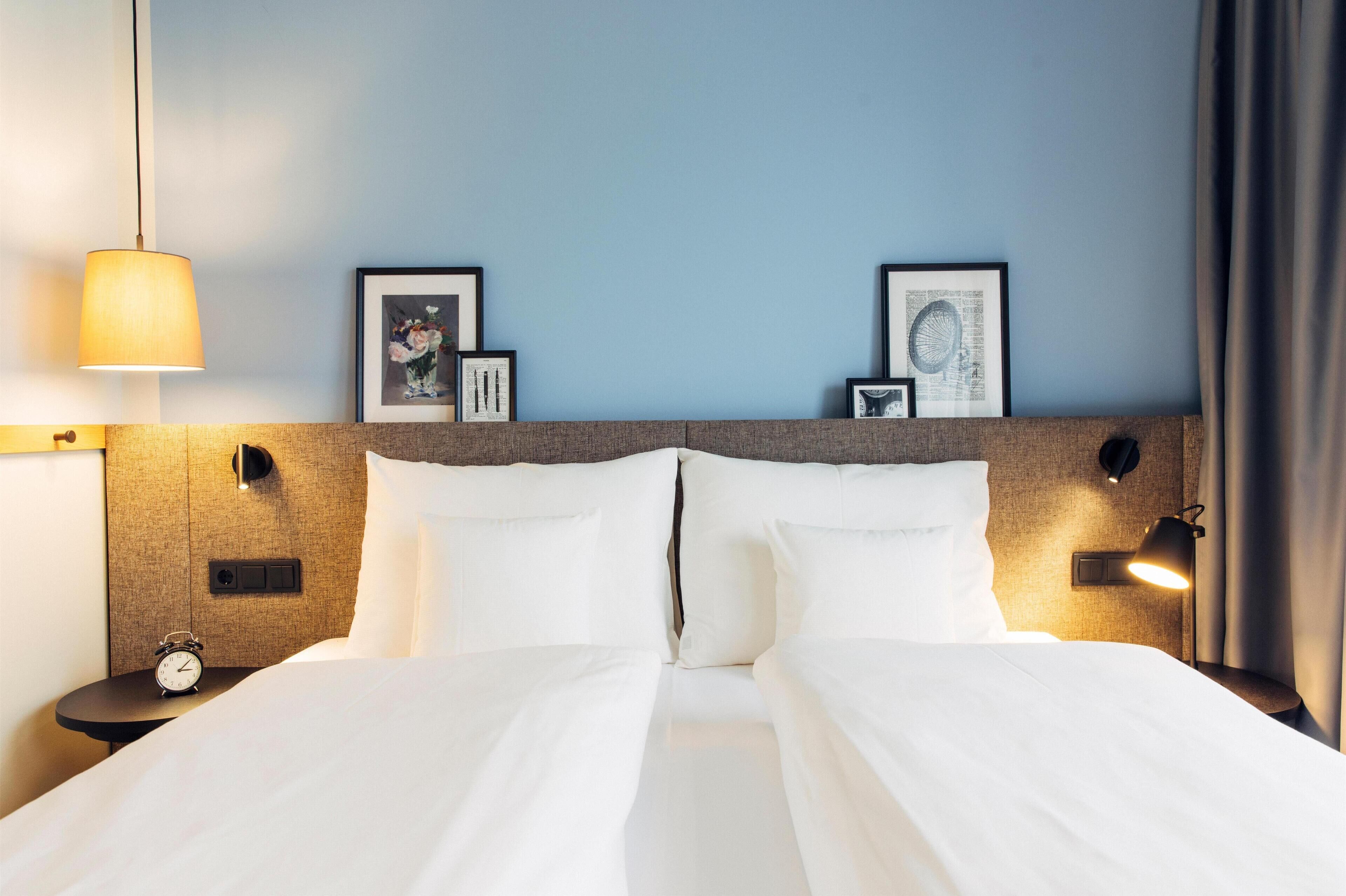 Una cama bien hecha con sábanas blancas, cabecera con luces fijadas a la pared y cuadros en la pared.