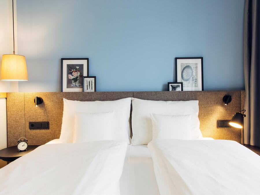 Ein ordentlich bezogenes Bett mit weißen Leinen, ein Kopfteil mit Wandleuchten und gerahmte Kunstwerke an der Wand.