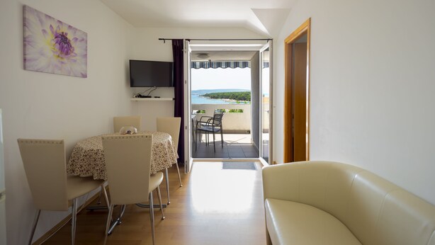 A room with a dining table, chairs, a TV mounted on the wall, and a view of the sea through an open door.