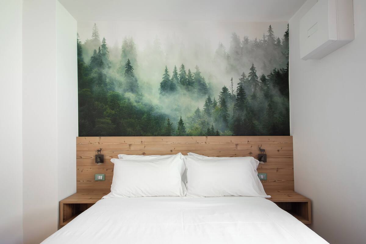 A bedroom with a large forest mural on the wall, a wooden headboard, and a neatly made bed with white linens.