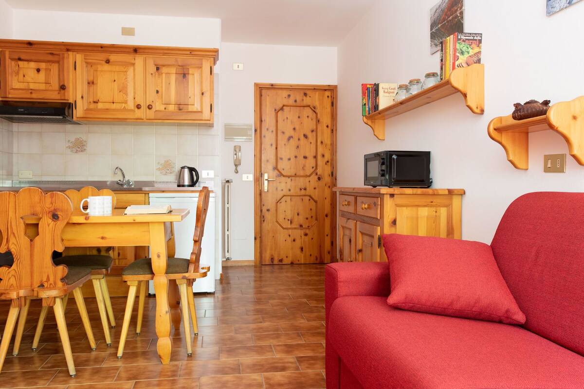 A compact living space with a wooden dining table, chairs, a red sofa, a kitchen area with wooden cabinets, and a microwave on a small countertop.