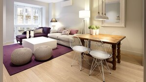 En moderne stue med et træbord, hvide stole med metalstel, en beige sofa og en lilla måtte.