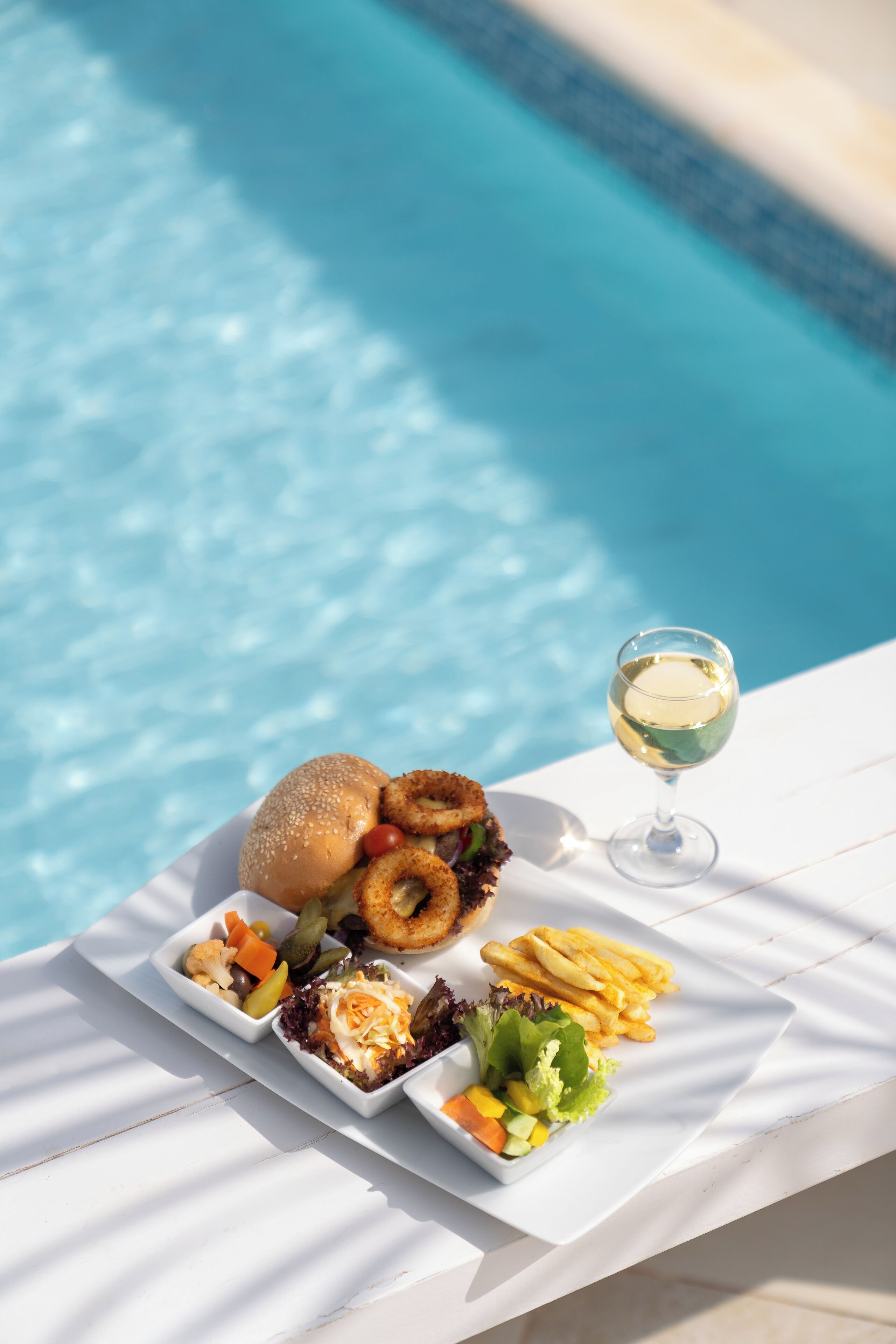 Una mesa de comedor con diversos alimentos y una copa de vino blanco junto a una piscina.