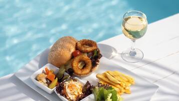 Una mesa de comedor con diversos alimentos y una copa de vino blanco junto a una piscina.
