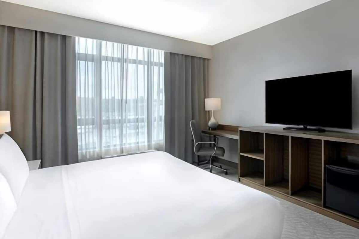 A hotel room with a large bed, a desk with a chair, a television, and a large window with curtains.