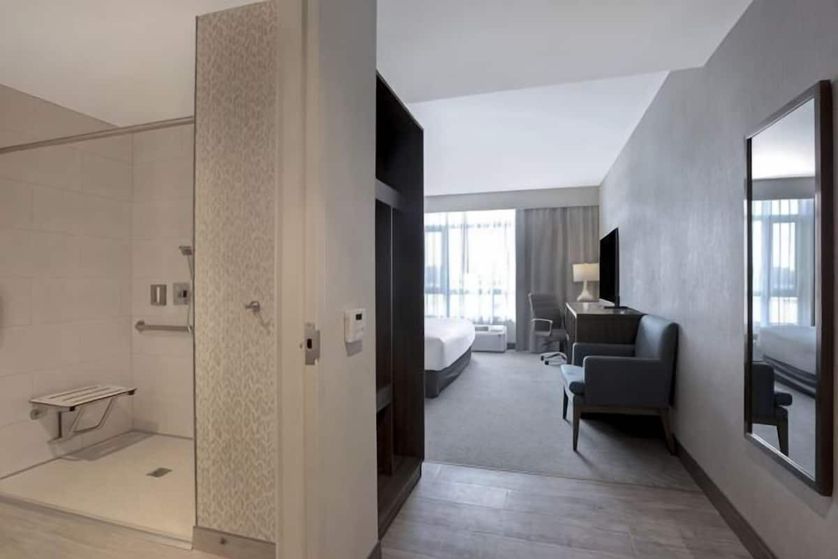 A hotel room with a shower, a bed, a desk, a chair, and a large window.