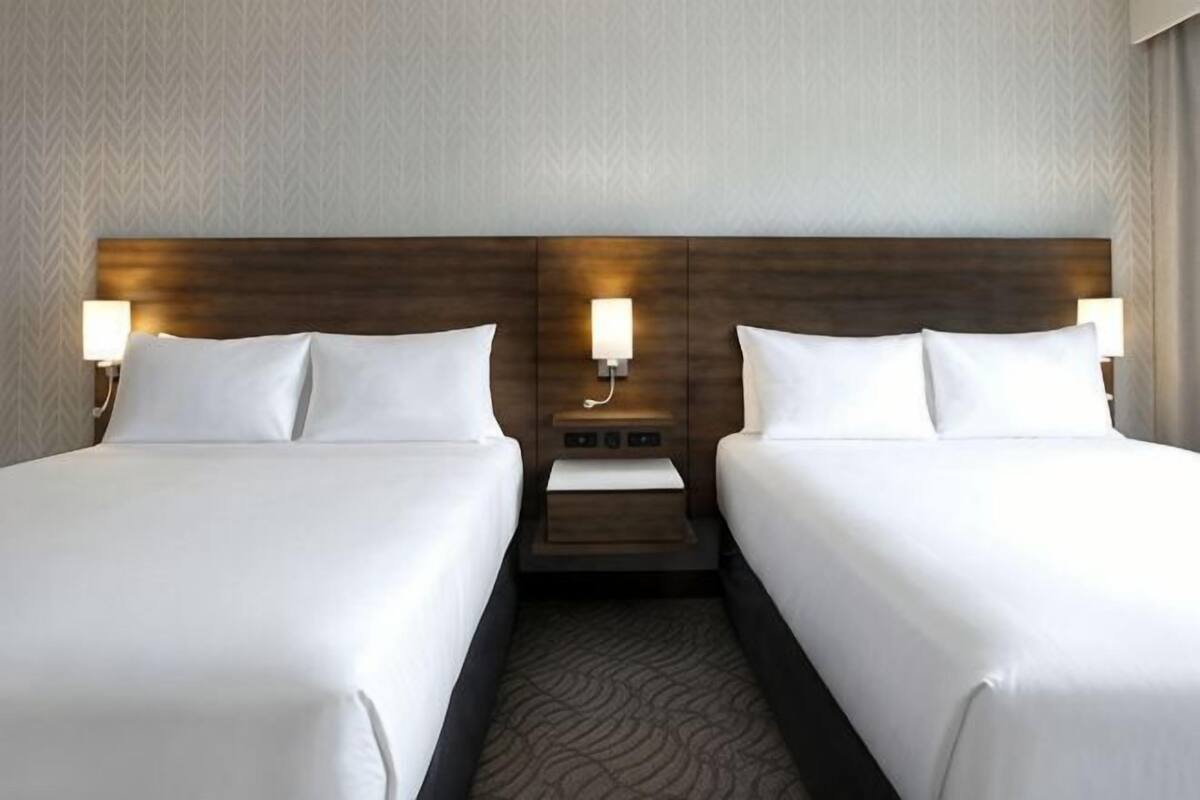 A hotel room with two beds, a nightstand, and wall-mounted lamps.