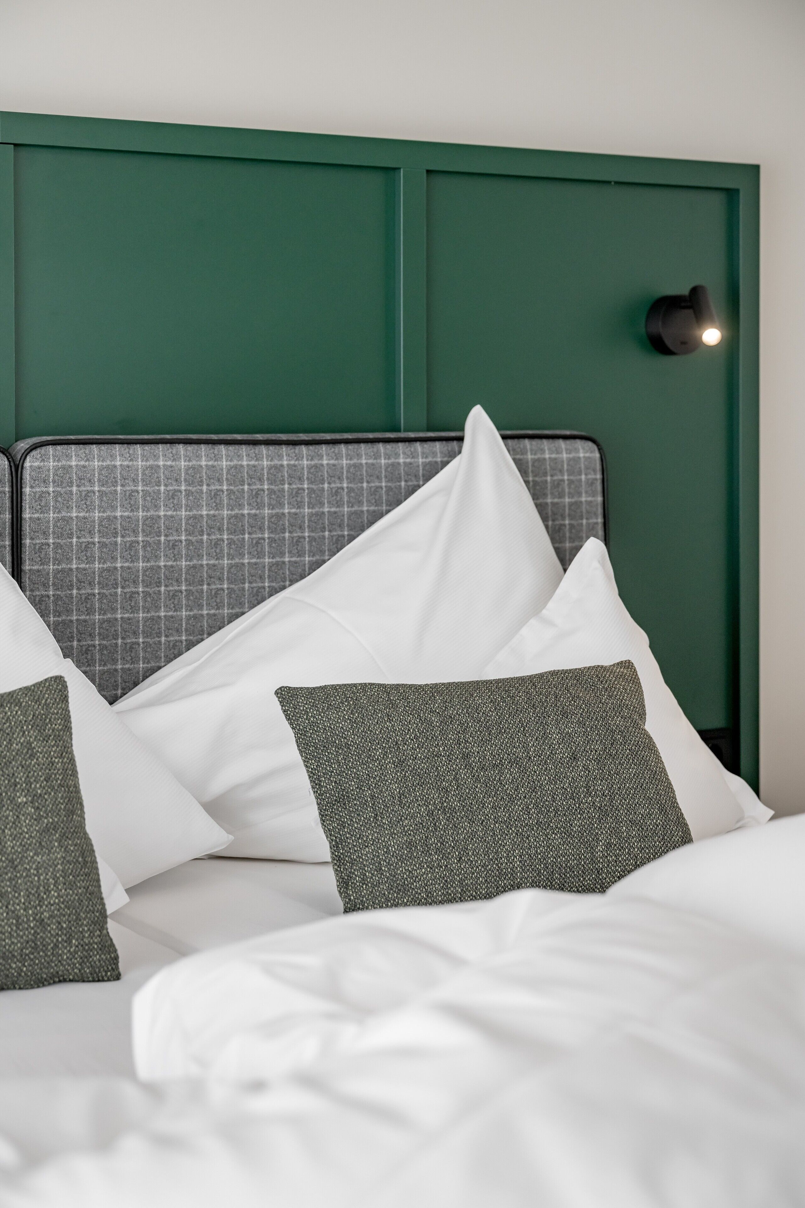 A neatly made bed with white and grey bedding, a green headboard, and a wall-mounted light fixture.