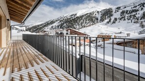 Un balcón de madera con barandilla de metal que da a un paisaje nevado de montañas y edificios.