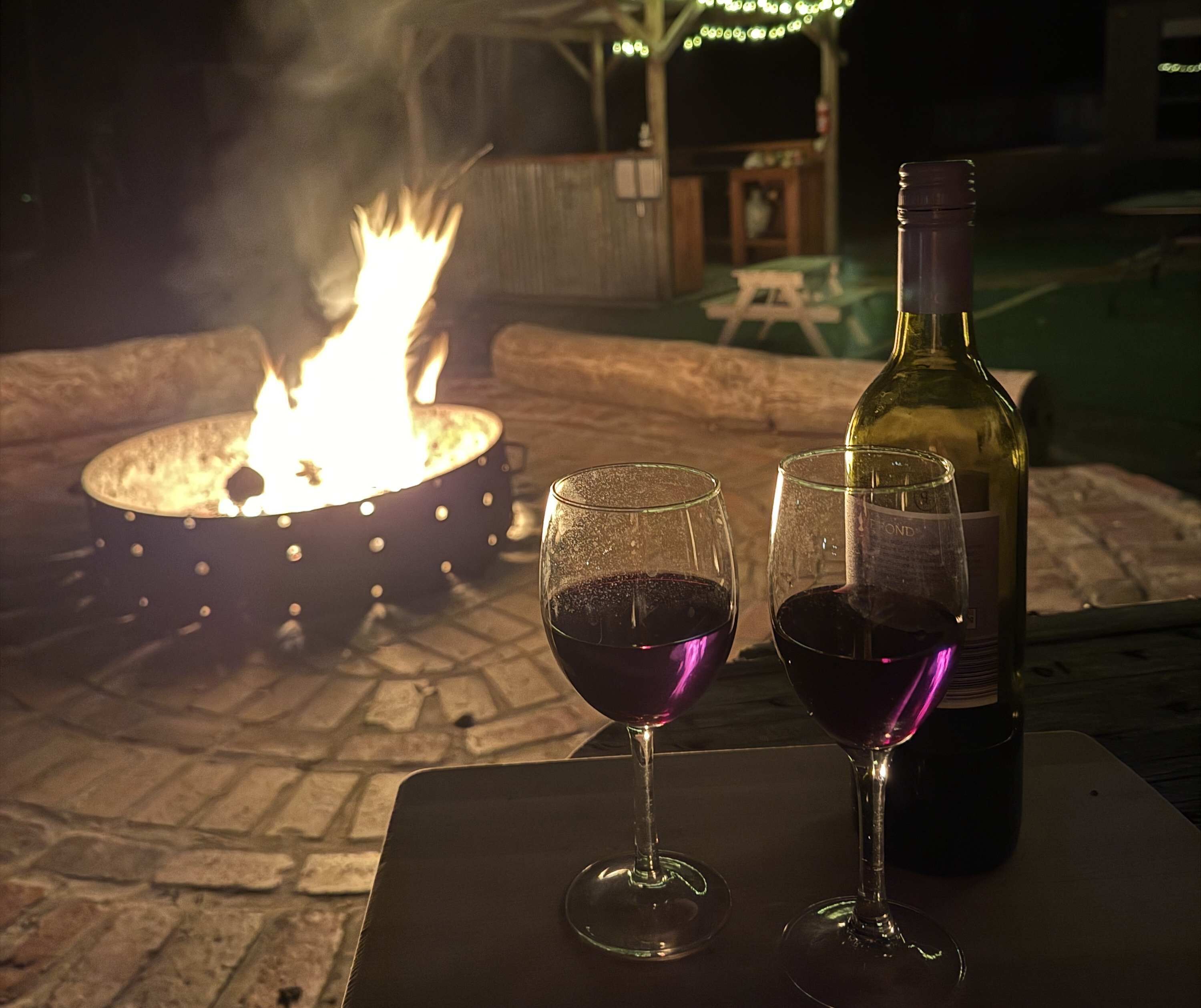 A table with two wine glasses and a bottle of wine, set against a backdrop of a fire pit.
