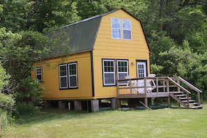 A yellow wooden house with a small deck and stairs, surrounded by trees.