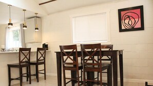 Un coin repas avec un sol carrelé de carreaux blancs, des chaises en bois, une table, des luminaires suspendus, une fenêtre avec des stores et une œuvre d’art abstraite encadrée accrochée au mur.